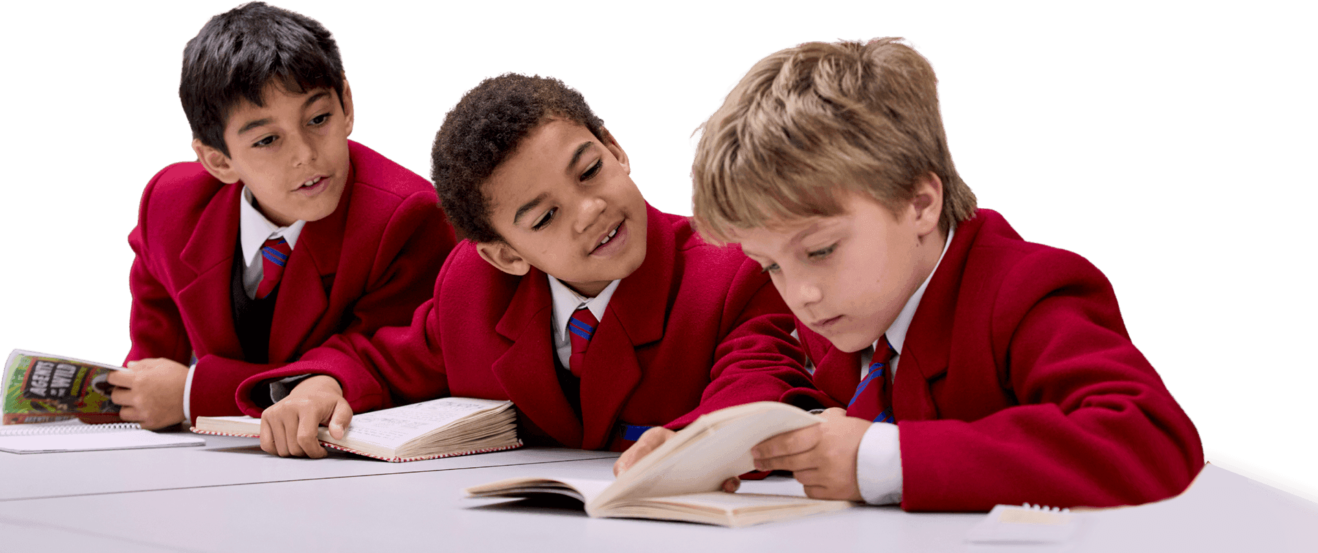 Three young boys in red blazers read books together at a classroom table.